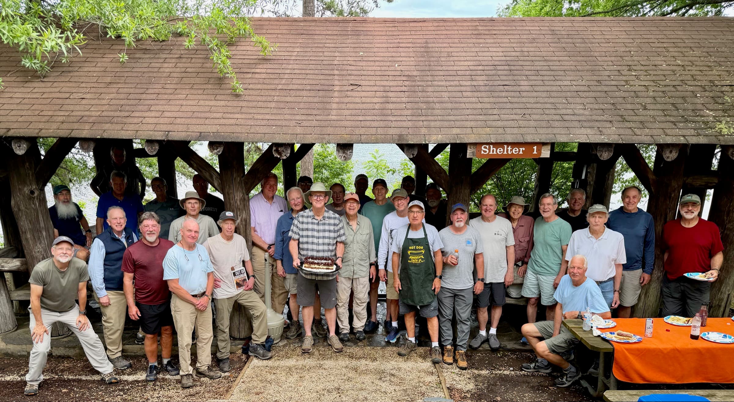 About 30 members of the BG Hilltrippers hiking group posing together under the timber-frame Shelter No. 1 at Holliday Lake State Park, one holding a cake.